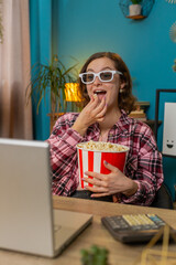 Happy businesswoman freelancer taking break from work wearing 3D glasses eating popcorn and watching movie on laptop. Smiling girl sitting at home office desk having snacks during break. Vertical