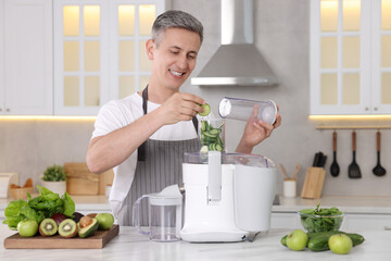 Wall Mural - Smiling man putting fresh cucumber into juicer at white marble table in kitchen