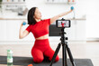 © Sofiia Shunkina - Woman in red sportswear recording fitness video at home on a smartphone. Fitness influencer demonstrating exercises with dumbbells for online audience. Healthy lifestyle and workout concept.