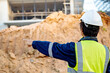 © Summer Paradive - Asian soil engineer in reflective green vest with helmet, earmuffs and goggles pointing at pile of laterite soil in front of unfinished building at site. Geotechnical engineering for construction base