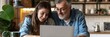 © nopommajun - A father and his teenage daughter sit together in a cozy kitchen, both focused and smiling as they look at a laptop screen, sharing a moment.