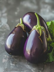 Poster - Three shiny eggplants with green leaves on a textured grey background. Suitable for rustic food photography, farm-to-table concepts, and vegetable features.