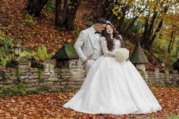 Wall Mural - A bride and groom are posing for a picture in a forest. The bride is wearing a white dress and the groom is wearing a suit. The couple is surrounded by trees and leaves, giving the image a natural