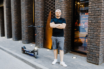 Young handsome man in n white baseball cap and casual clothies standing near electric scooter on the street.