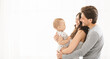 © Prostock-studio - A young couple smiles as they hold their baby in their arms. They are standing in front of a white background, and the baby is looking off to the side, copy space