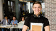 © Prostock-studio - A cheerful young man student stands in the foreground with a clipboard in his hand and headphones around his neck, flashing a bright smile