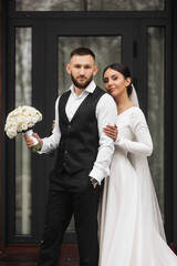 Wall Mural - A bride and groom are posing for a picture in front of a black door. The bride is wearing a white dress and the groom is wearing a black suit. They are holding a bouquet and a vase, respectively