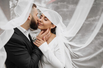 Wall Mural - A bride and groom are kissing each other under a white veil. The bride is wearing a white dress and the groom is wearing a black suit