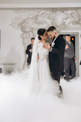 Wall Mural - A bride and groom are dancing in a foggy room. The bride is wearing a white dress and the groom is wearing a black suit