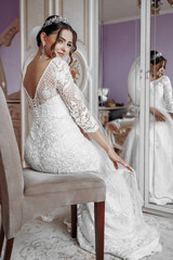 Wall Mural - A woman in a white wedding dress sits on a chair in front of a mirror. She is smiling and looking at her reflection. Concept of happiness and celebration, as the woman is getting ready for her wedding