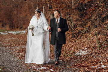 Wall Mural - A bride and groom are walking down a path in the woods. The bride is wearing a white dress and a fur stole, while the groom is wearing a suit. They are holding hands