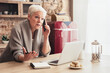 © Prostock-studio - A senior woman with white hair sits at a kitchen table talking on a mobile phone while gesturing with her other hand. She is sitting in front of a laptop computer and a notebook.