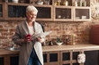 © Prostock-studio - A senior woman stands in her kitchen, wearing a gray cardigan and jeans. She holds a red mug in one hand and reads a newspaper with the other, copy space