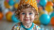 © svastix - A cheerful toddler with curly hair, wearing a vibrant polka-dotted orange hat, smiles charmingly in front of colorful balloons in the background.