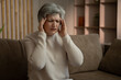 © Grustock - An elderly woman is sitting on the couch in her living room. She has a headache and is clearly uncomfortable
