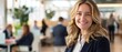 © mikeosphoto - A smiling young blonde woman in a business suit stands in a bright, modern office. The background includes blurred colleagues and natural light