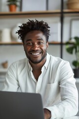 Wall Mural - Attractive African American businessman sitting at his desk with a laptop, smiling and looking at the camera, wearing a white shirt, with short black hair and beard, against a light background.