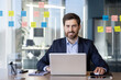 © Liubomir - Confident businessman in suit smiling while working on laptop in modern office environment. Organized workspace with colorful sticky notes and natural light. Productive work atmosphere and success.