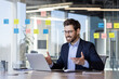 © Liubomir - Businessman engaged in virtual meeting using laptop in modern office. Colorful post-its on glass walls indicate brainstorming planning session. Professional setting with confident, happy expression.