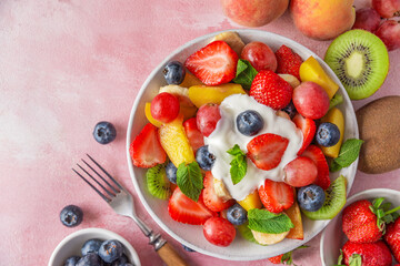 Wall Mural - Healthy fresh fruit salad with yogurt in a bowl with fork on pink background. Top view with copy space. Summer healthy food for breakfast. Mixed tropical fruits berries for vegan diet lunch