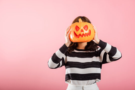A portrait of a joyful Asian woman holding model pumpkins on her head in a Halloween-themed studio shot. She's all smiles with ghost pumpkins on a pink background.