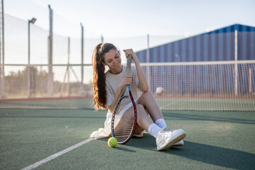 young woman sits on a tennis court, smiling and holding a tennis racket. The sun shines brightly, and the court is green