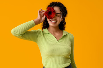 Wall Mural - Beautiful young African-American woman with red gerbera flower on yellow background. International Women's Day