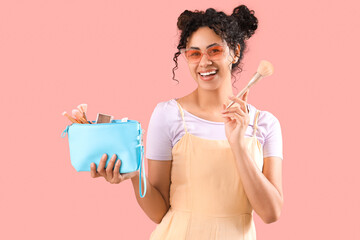Wall Mural - Beautiful young happy African-American woman with cosmetic bag and makeup brushes on pink background