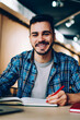 © BullRun - Portrait of cheerful male student enjoying learning in coworking office using laptop computer for research,happy freelancer looking at camera during making project for remote job making notes