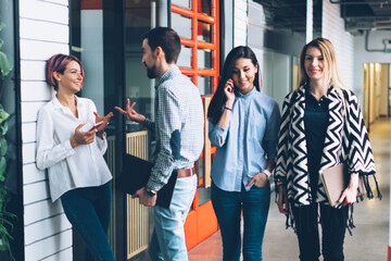 Wall Mural - Attractive cheerful woman talking with man colleague during downloading application on smartphone connected to internet while hipster girls passing along and communicating using modern cellular