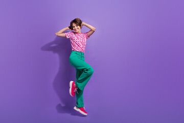 Wall Mural - Full length photo of gorgeous pretty girl wear stylish t-shirt green pants flying hold hands on head isolated on purple color background
