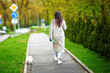 © Tricky Shark - Woman walking her white pomeranian dog on leash in city park, small pet and owner. Female in coat strolls with her white pomeranian on leash along the city park sidewalk