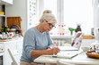 © pikselstock - Senior woman with laptop and notebook sitting at the table in the kitchen