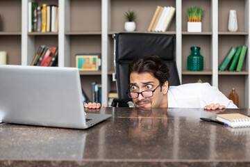 Wall Mural - Shocked businessman hiding behind desktop and looking at laptop in the office. Business crisis and bankruptcy concept