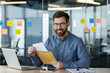 © Tetiana - Portrait of a smiling young man in glasses and a blue shirt sitting at a desk in the office, holding an open letter in a yellow envelope, looking at the camera