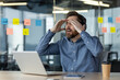 © Tetiana - Shocked young man working in the office at the laptop, sitting at the desk and holding his head with his hands in frustration