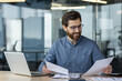 © Tetiana - Smiling young man businessman working in office with documents and laptop, holding and reviewing papers
