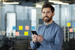 © Tetiana - Portrait of a young smiling man in a blue shirt and with a beard standing in the office on a transparent whiteboard with colored stickers, holding a mobile phone and looking at the camera
