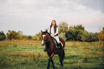 young beautiful blonde woman jockey rides a brown horse in a meadow at sunset in summer. preparing f