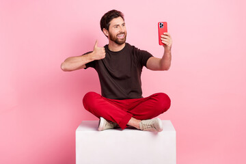 Full size photo of nice man dressed brown t-shirt sit on cube show thumb up make selfie on smartphone isolated on pink color background