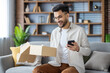 © Tetiana - Muslim smiling young man in casual clothes and glasses is sitting on the sofa at home, holding a mobile phone and looking into an open box of a package he received by mail