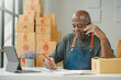 © amnaj - Man wearing apron at desk with tablet, on phone, writing in notebook. Stack of parcels behind him, engaged in small business activities.
