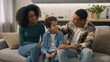 © Yuliia - African American happy family portrait couple man father male dad mom woman mother female sitting indoor couch talking speak cuddle stroking head little boy child son kid parent tenderness living room