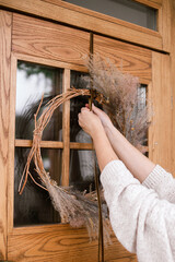 Woman decorating front door with stylish autumn wreath. Rustic wreath with dried grass, herbs and ribbon in hands. Fall decor of house entrance. Thanksgiving and Halloween