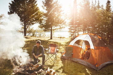 Couple, campfire and wood with smoke in nature by tent on grass field for outdoor vacation, holiday or weekend. Man preparing fire by woman for camping leisure, summer getaway or sunshine at forest