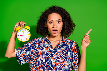 Wall Mural - Photo of lovely young lady hold look alarm clock point empty space dressed stylish colorful garment isolated on green color background