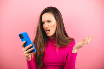 Wall Mural - Mad and shock young brunette beautiful woman using mobile phone isolated on pink background. Yelling unhappy woman hold mobile phone. Disappointed sad upset lady horrified impressed news. WTF. Oh no.