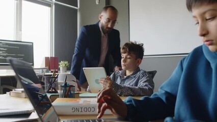 Medium close-up of Indian boy typing on laptop, Caucasian male student using on tablet computer, while working on programming project in classroom, and teacher walking around, checking their progress
