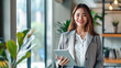 © mimi - A successful young businesswoman standing confidently with a tablet, surrounded by greenery in a modern office, exuding professionalism and a dynamic approach to work.