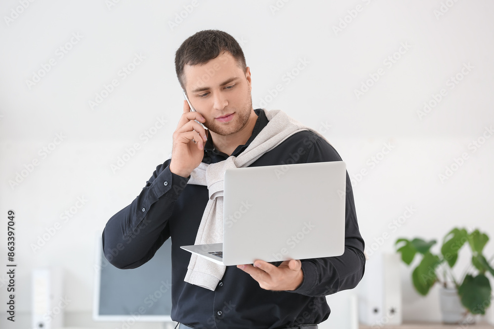 Young businessman with laptop talking by mobile phone in office